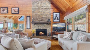 Una sala de estar acogedora con chimenea de piedra, techo de madera y amplios ventanales que ofrecen vistas al exterior.