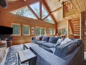 Interior de una cabaña de troncos con un amplio sofá modular, un televisor de pantalla plana y amplias ventanas que dan vistas al bosque circundante.