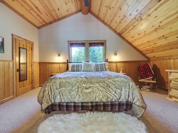 Un dormitorio acogedor con techo de madera, una cama grande con manta mullida, dos lámparas de noche y una ventana con cortinas.