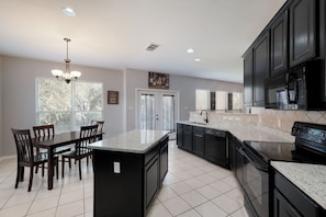 A modern kitchen with dark cabinetry, a large island, and a dining area with chairs. - SeaWorld 3 Minutes. Lackland AFB Nearby (San Antonio)