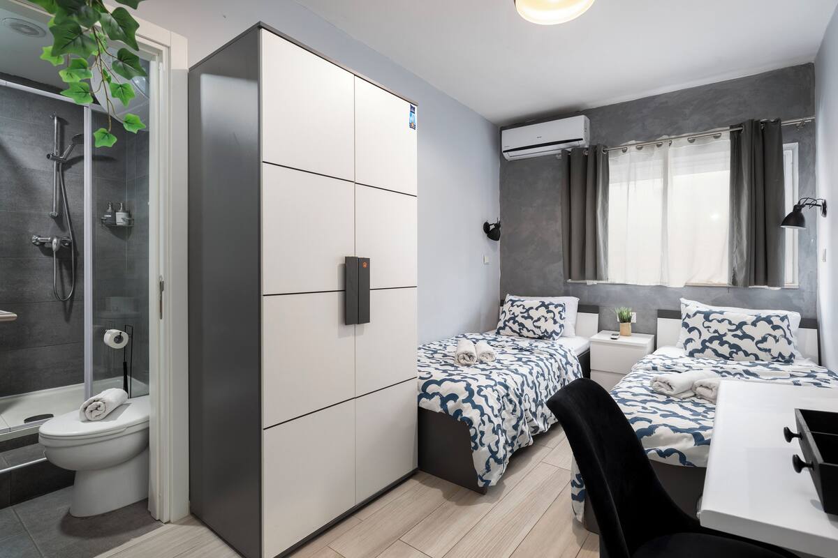 Une chambre d’hôtel compacte, dotée d’une douche, de deux lits, d’un petit bureau et d’une fenêtre avec des rideaux.