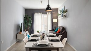 A modern dining area with a white table set for four, a black pendant light, and a view of the living room with a sofa and cushions. - Modern Ensuite Rooms Near Balluta Bay (St. Julian's)
