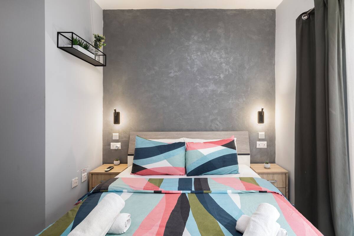 Une chambre moderne avec un grand lit, deux tables de chevet, une étagère murale agrémentée de plantes et un mur gris à texture.