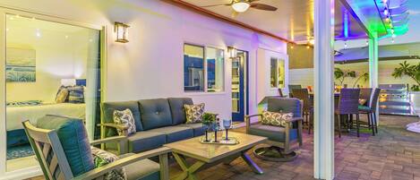 A modern lounge area with a sofa, chairs, and a table. There is a ceiling fan, a window with a view, and a door leading outside.