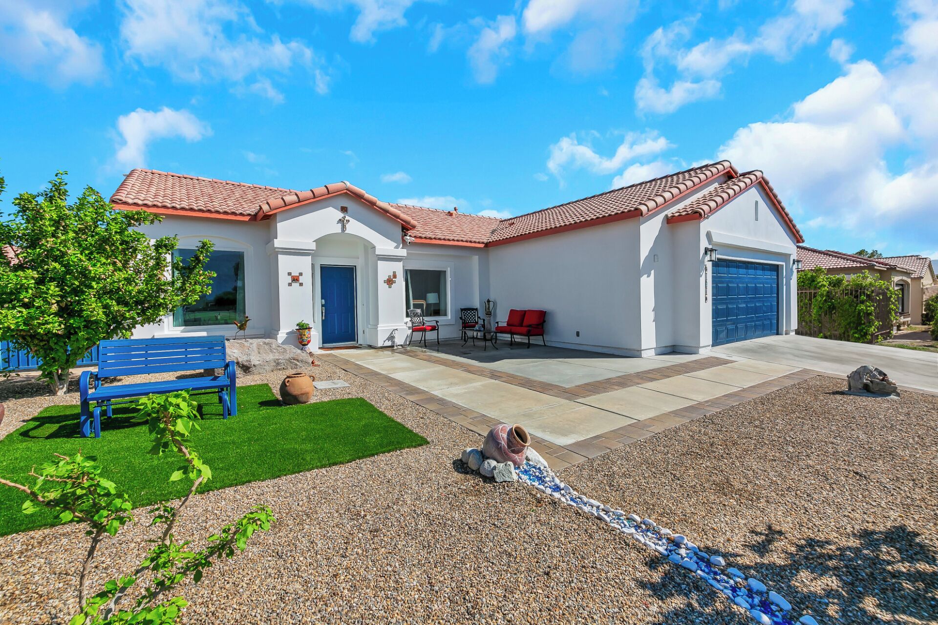 A two-story house with a red-tiled roof, a blue garage door, and a front yard with a bench, a red chair, and a decorative stone pathway.