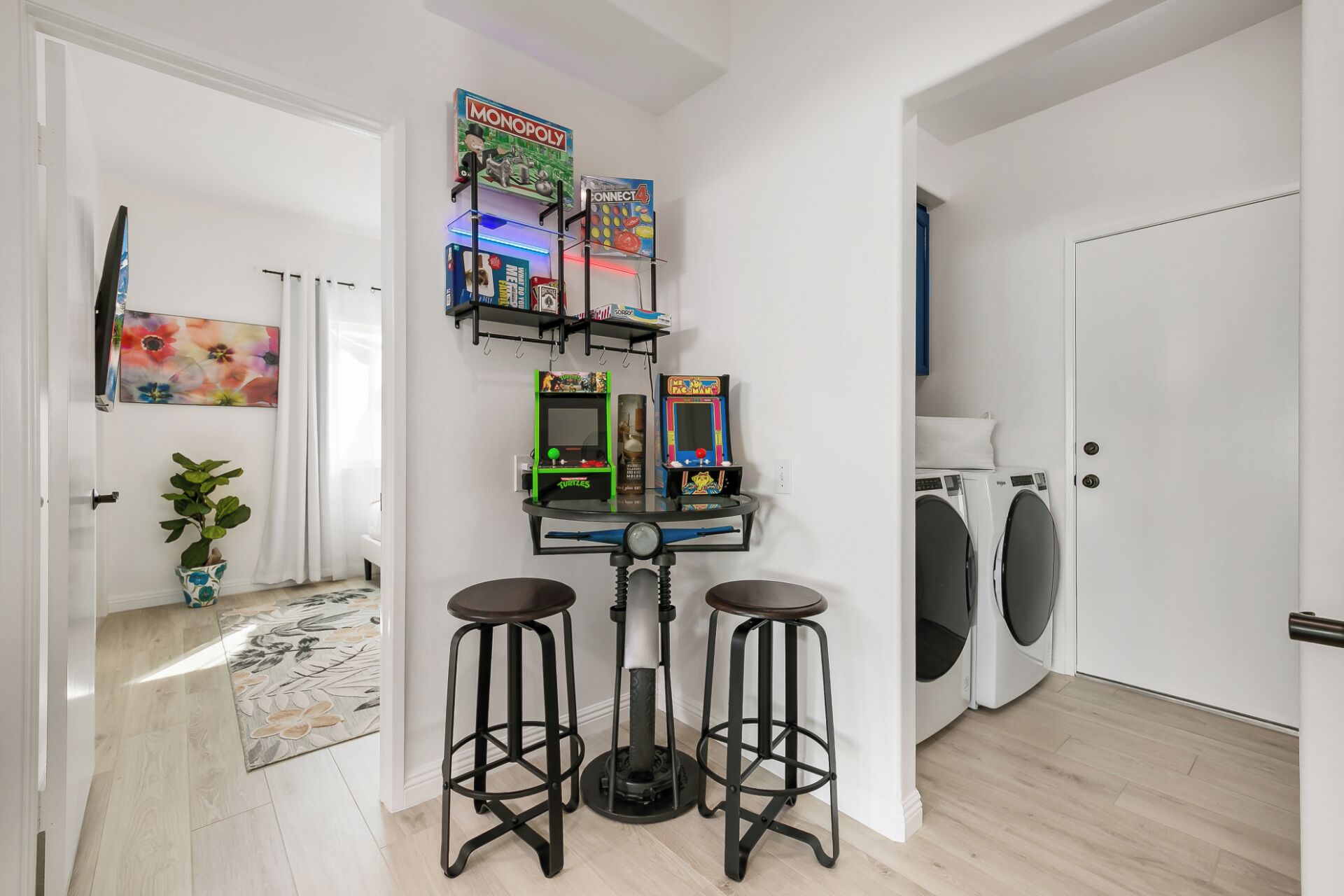 A room with a washing machine, two bar stools, a shelf with games, and a door.