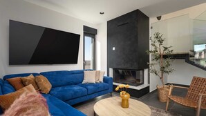 Ein modernes Wohnzimmer mit einem blauen Sofa, einem runden Couchtisch mit einer Vase, einem Flachbildfernseher, einem Kamin und Blick ins Freie.