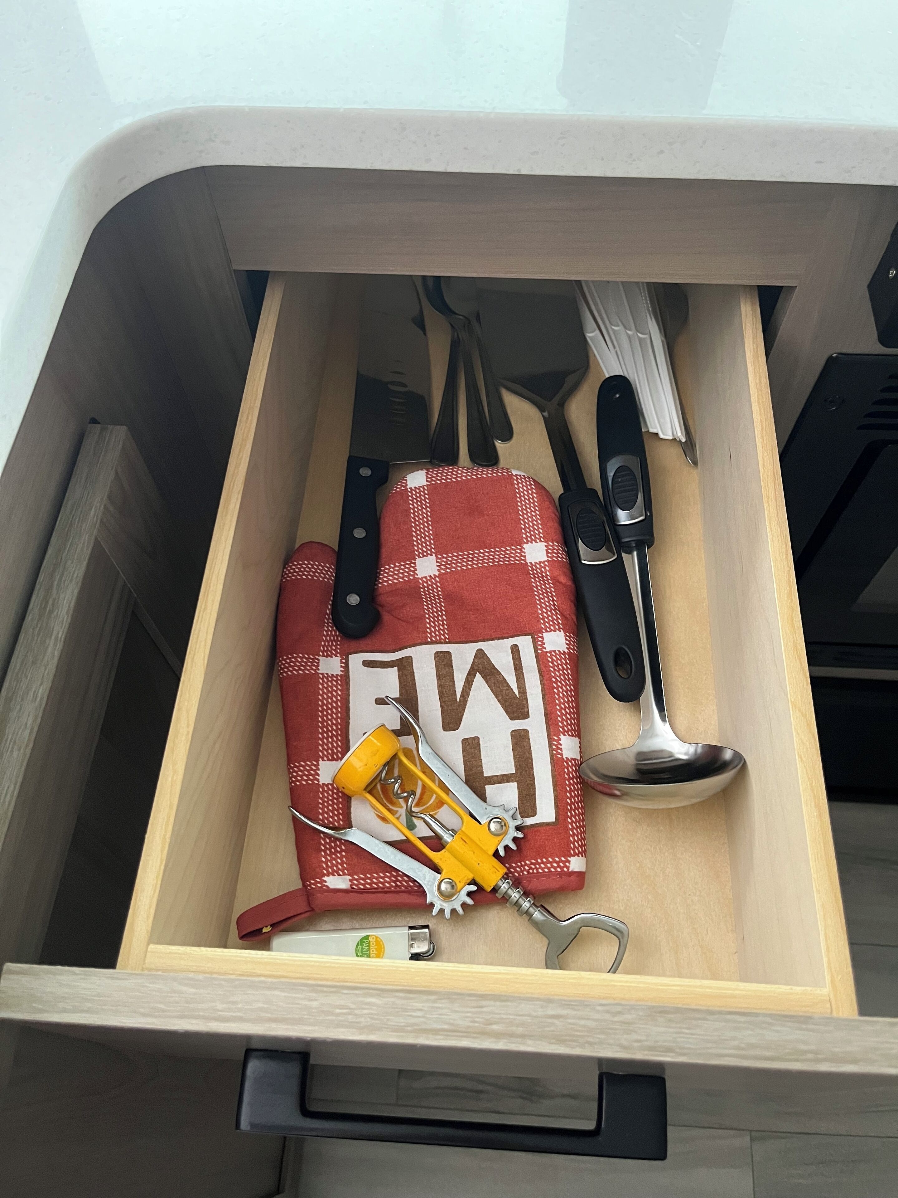 A drawer with kitchen utensils including a red and white checkered oven mitt, a knife, a spoon, and a can opener.