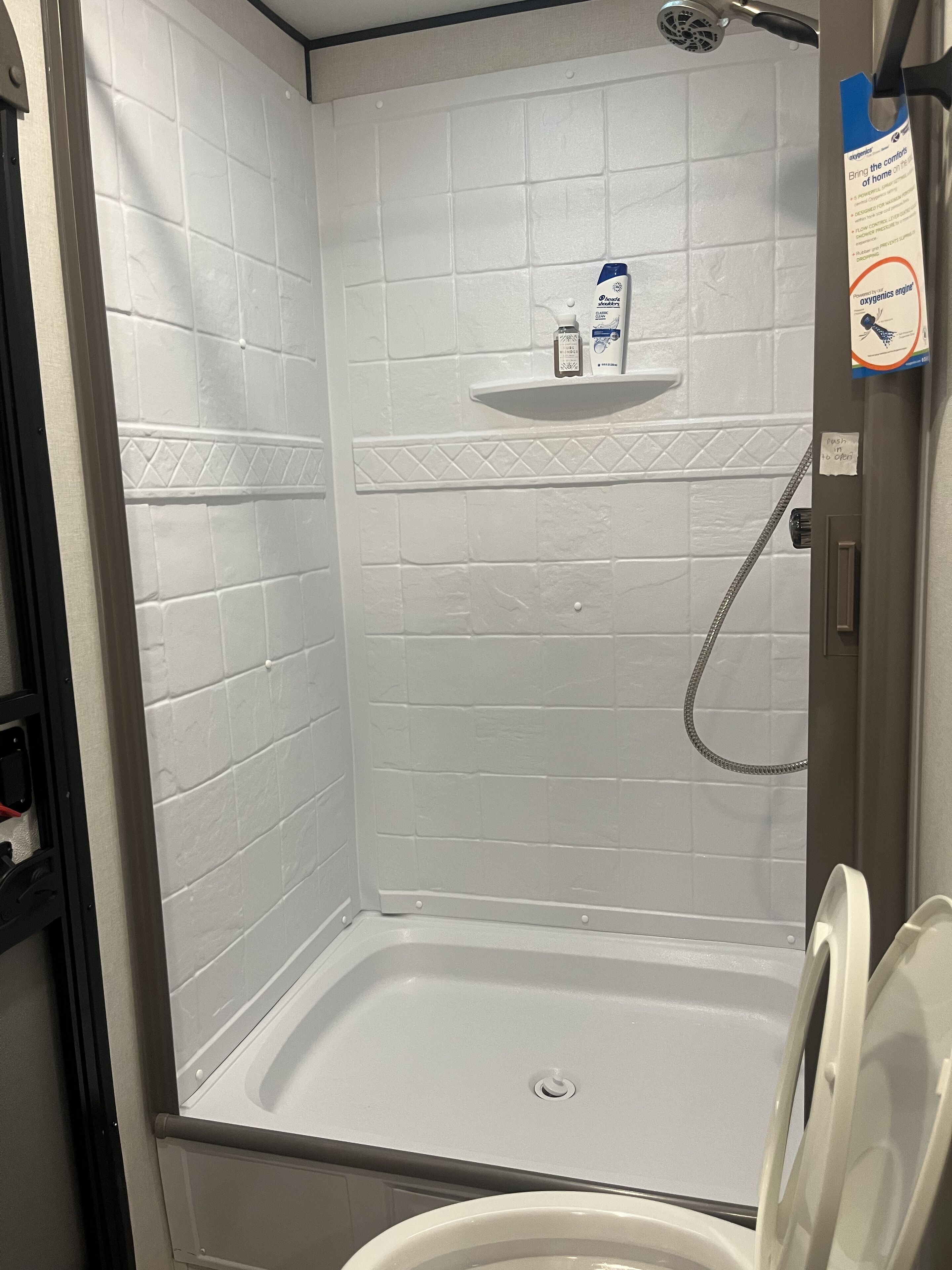 A shower stall with a white tiled wall, a shelf with toiletries, and a handrail.