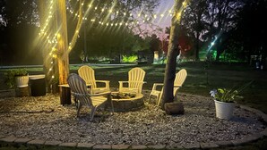 Un espacio al aire libre con sillas Adirondack, un hogar de piedra y luces colgantes.