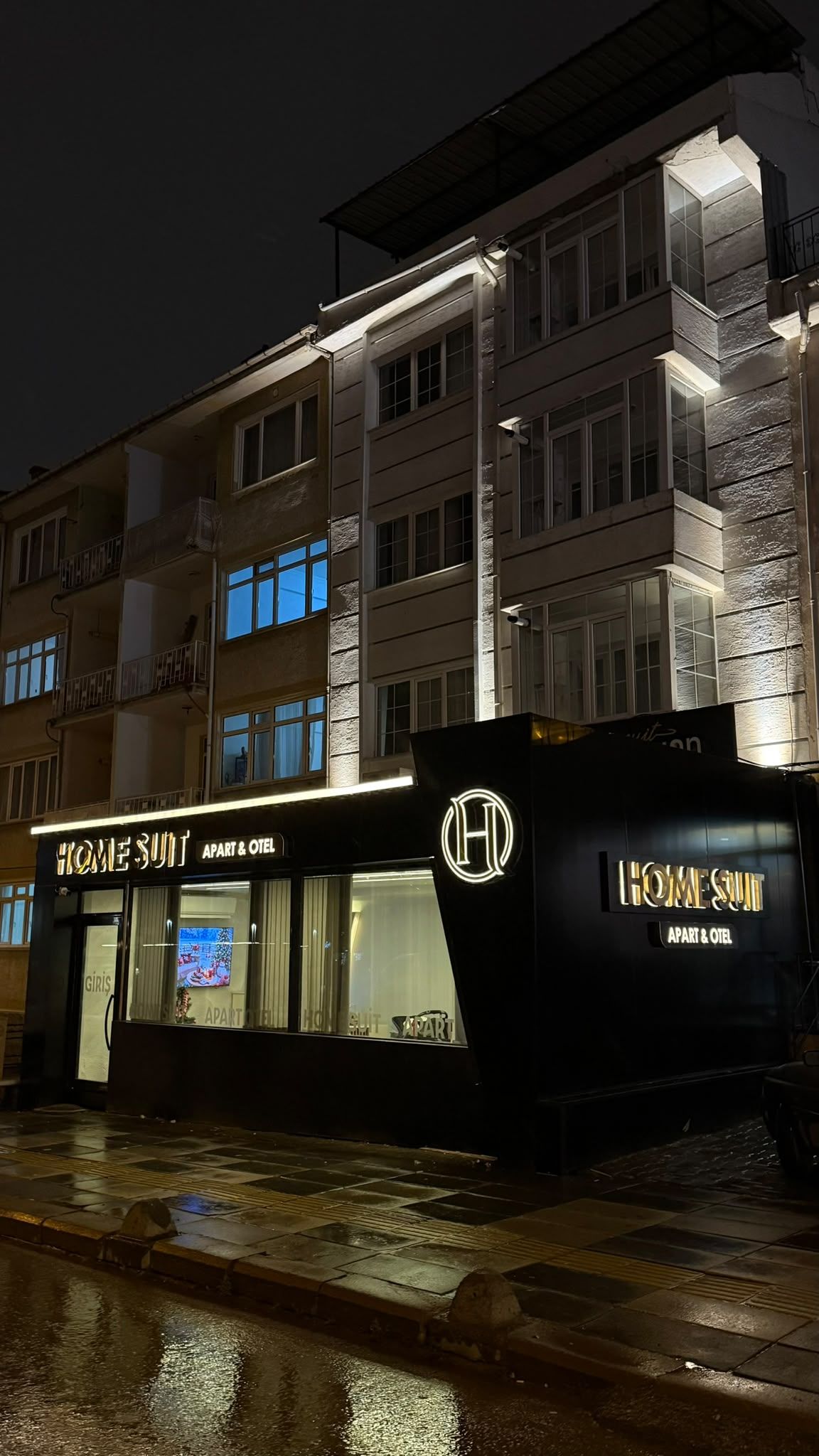 Un edificio con un letrero que dice 'HOME SUIT APART & OTEI' y un logotipo con una 'H' mayúscula.