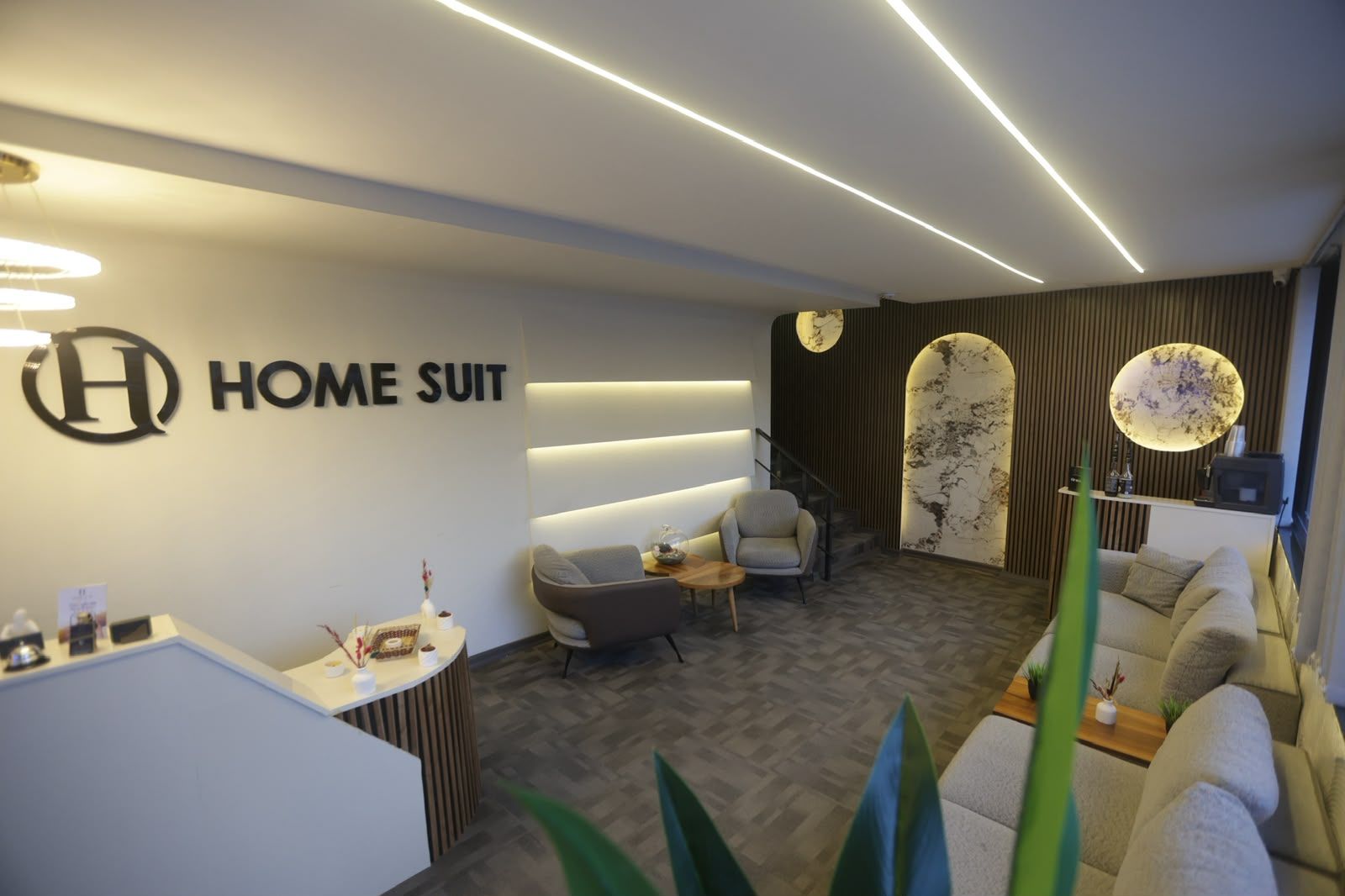 Ein moderner Empfangsbereich mit einer Rezeption, Sitzgelegenheiten und einem an der Wand befestigten Schild mit der Aufschrift 'HOME SUIT'.