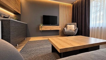 Una sala de estar moderna con una mesa de centro de madera, un sofá gris, un televisor montado en la pared y un ventanal con cortinas.