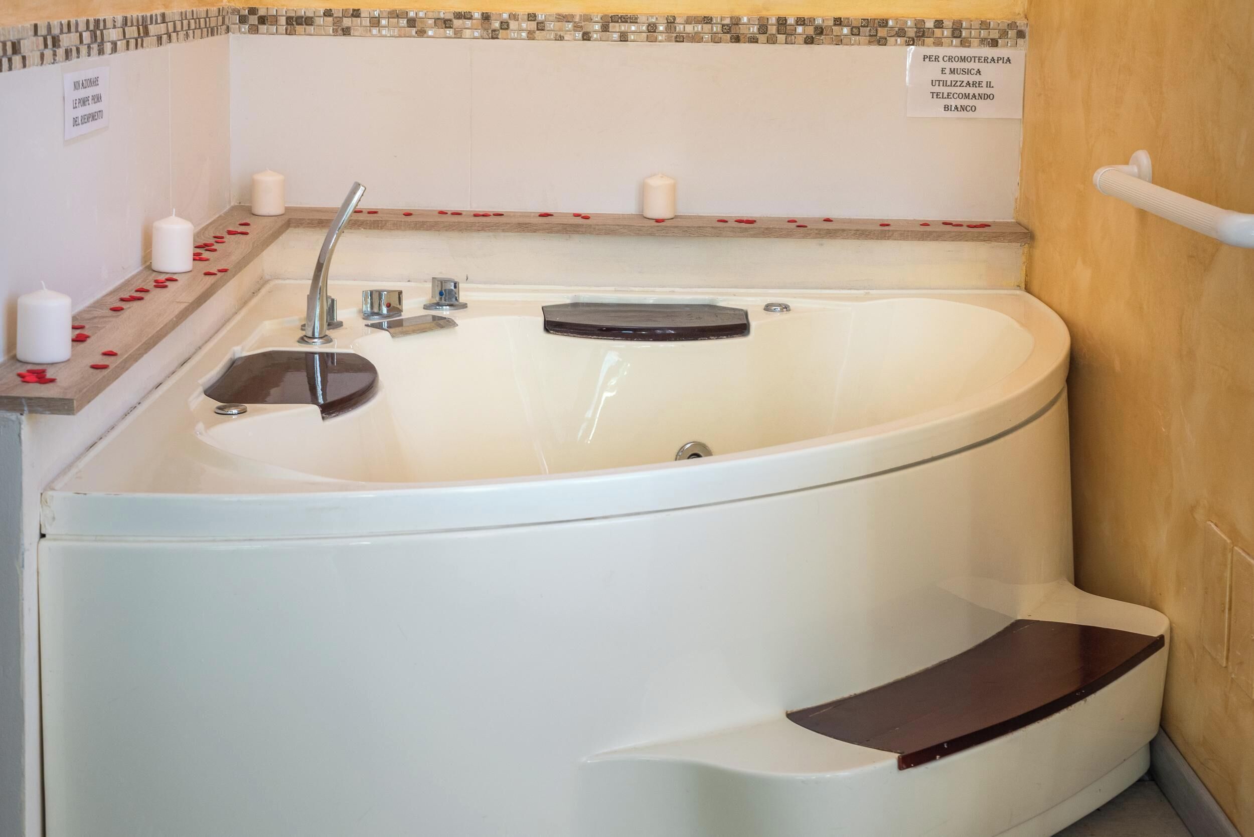 A large, white freestanding bathtub with a wooden tray and a built-in seat, surrounded by a tiled wall with a decorative border.