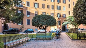 A paved walkway with benches, parked cars, and a multi-story building with balconies. - Roma Design San Giovanni Colosseo area (Rome)