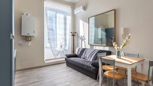 A modern living room with a grey sofa, wooden dining table, and chairs. There is a large window with sheer curtains, a mounted air conditioner, and a decorative vase with flowers. - Roma Design San Giovanni Colosseo area (Rome)