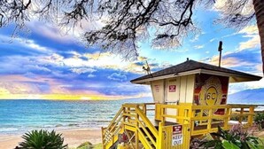 A lifeguard tower with a 'KEEP OUT' sign, located near a beach with a view of the ocean and a sunset.