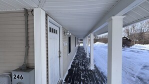 A covered porch with a snow-covered pathway leading to a wooden cabin. - Island Pond Apartment on VAST Trail – 2BR, 1BA (Brighton)