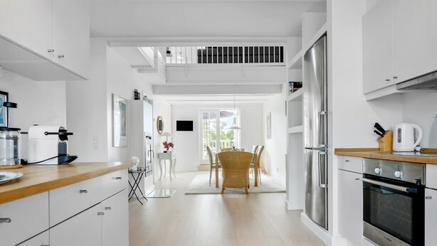 A modern kitchen with white cabinetry, a dining area with a wicker table, and a view of the outdoors through a large window. - Awesome home in Gråsten with sauna (Gråsten)