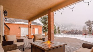 A covered outdoor patio with a fire pit, wicker furniture, and a brick building in the background. - Spacious 6-Bedroom Mountain Home –Minutes from Park City’s Year-Round Adventures (Kamas)