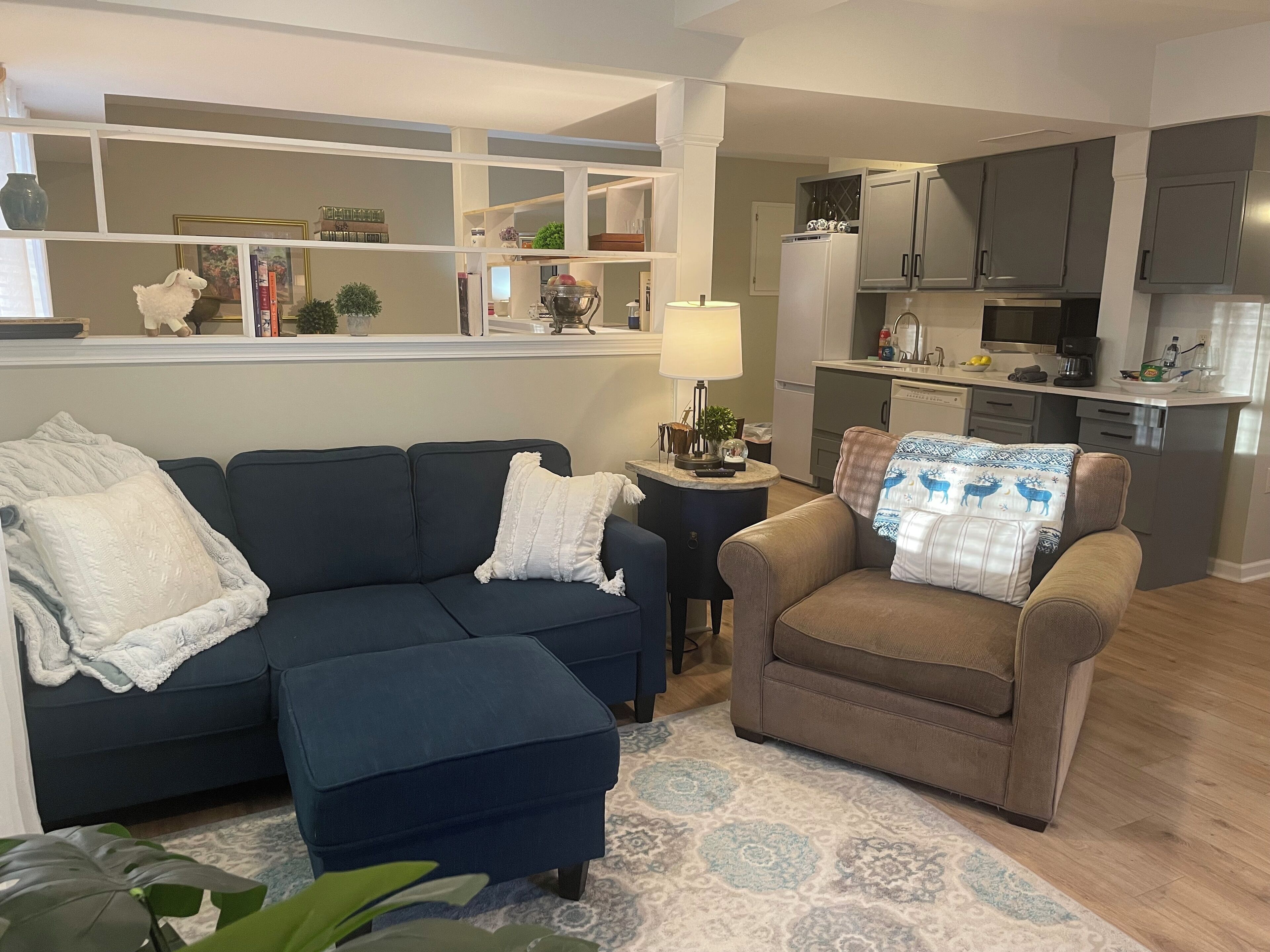 Ein Wohnzimmer mit einem blauen Sofa, einem braunen Sessel und einem gemusterten Teppich. Ein kleiner Beistelltisch mit einer Lampe und Dekorationselementen ist ebenfalls vorhanden. Im Hintergrund ist eine Küchenzeile mit modernen Geräten und Schränken zu sehen.