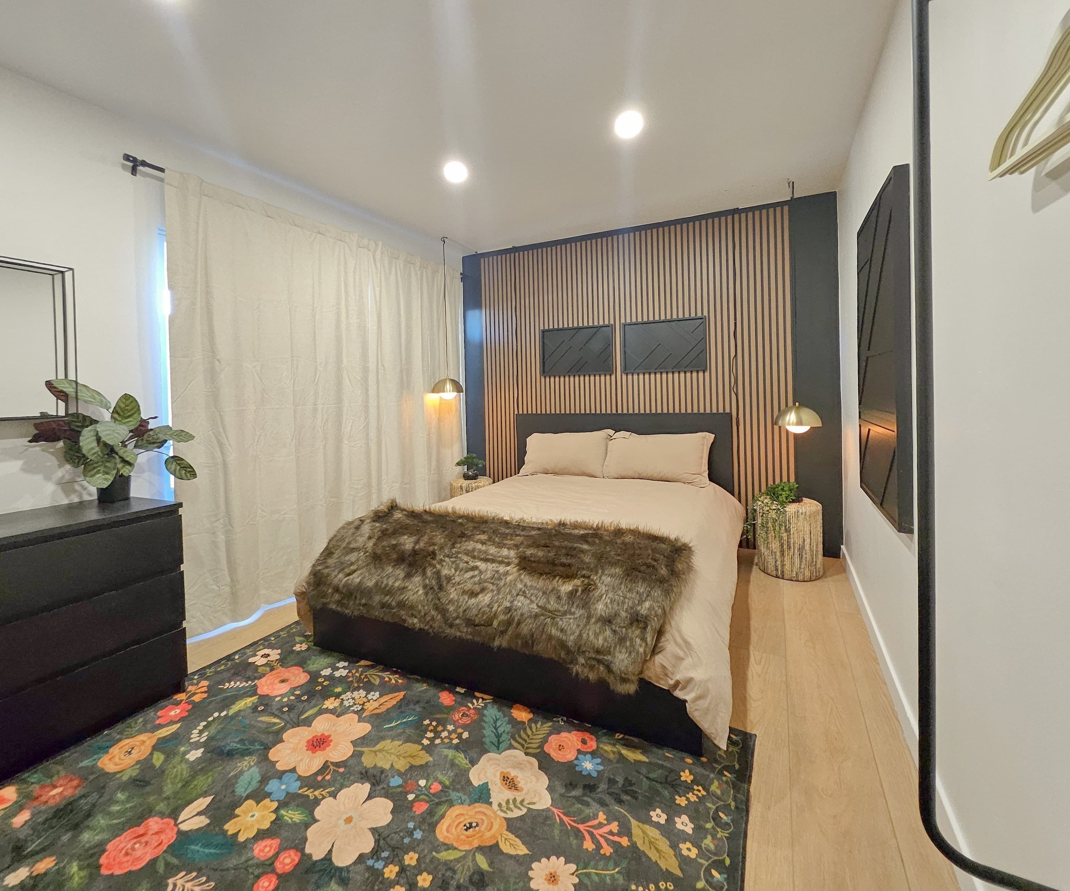Un dormitorio moderno con cama, cómoda, alfombra estampada y televisor montado en la pared.