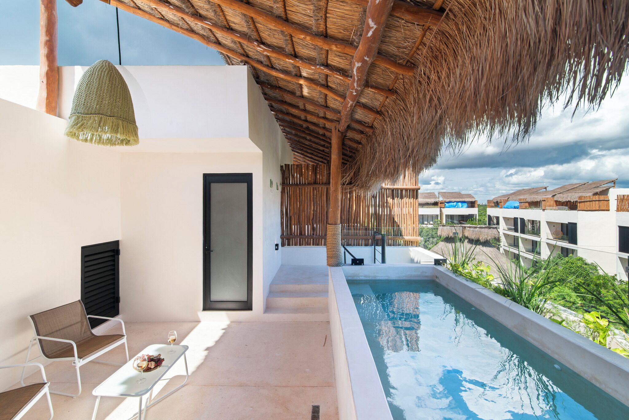 A poolside area with a thatched roof, a seating area with a table, and a view of other buildings.