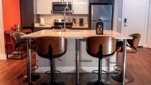 A modern kitchen with white cabinets, a stainless steel refrigerator, a microwave, and a coffee maker. There are two bar stools with wooden seats and black metal legs. - 2 Bedroom Buckhead Suite (Atlanta)