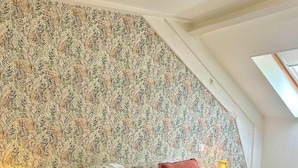 A bedroom with a sloped ceiling, floral wallpaper, a bed with two pillows, a bedside lamp, and a nightstand.
