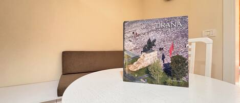 Une table ronde sur laquelle repose un livre intitulé « Tirana vue du ciel », un canapé marron, une chaise blanche, une horloge murale et un interrupteur.
