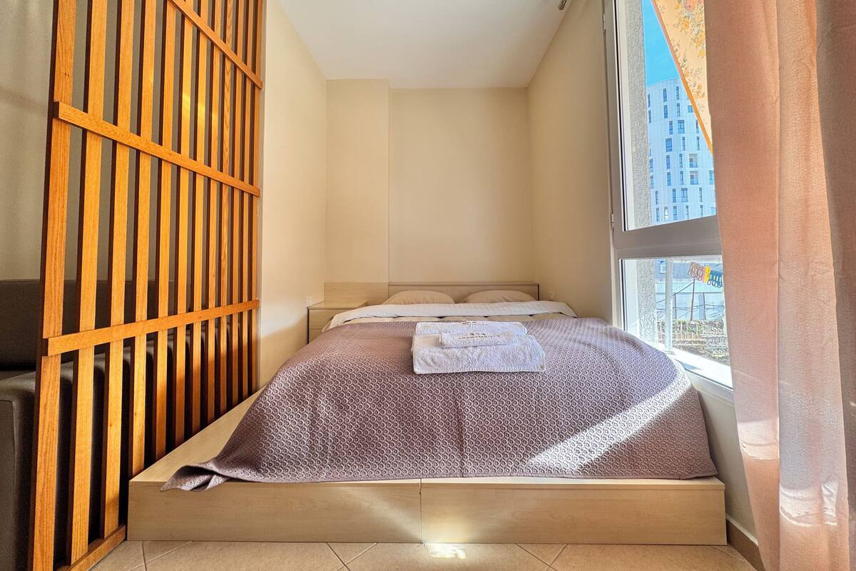 A small, modern bedroom with a bed, a wooden partition, and a window with a city view.