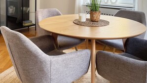 Een eetgedeelte met een ronde tafel, gestoffeerde grijze stoelen, een gele hanglamp, een vaas met gedroogde planten en een lijst met een afbeelding aan de muur.