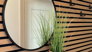 Een spiegel met een ronde lijst, een gevlochten mand met groene planten, en een houten lambrisering met haken.