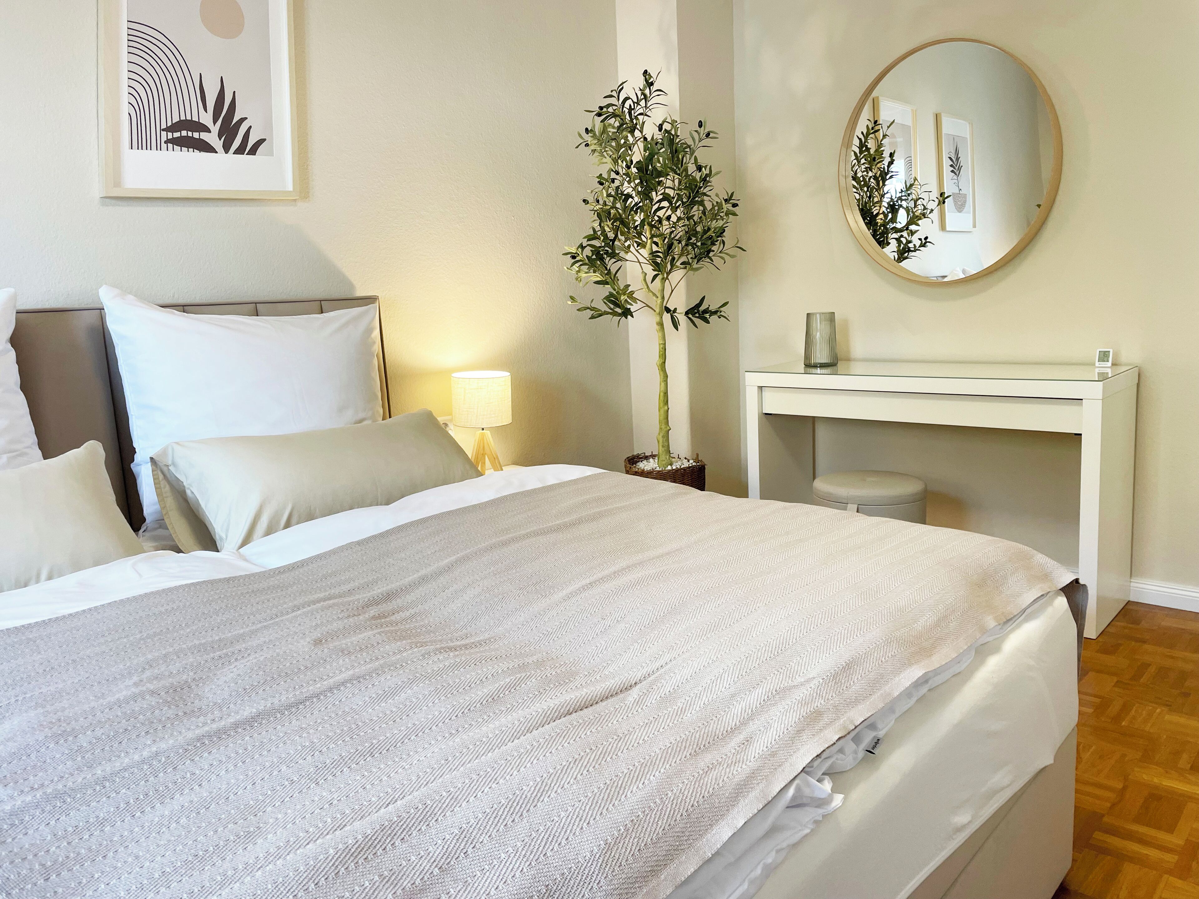 A neatly made bed with white linens, a bedside lamp, a small potted plant, a round mirror, and a white desk with a lamp.