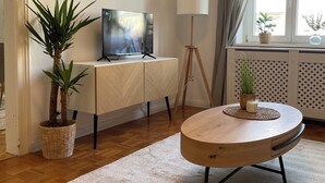 Een woonkamer met een televisie op een houten kast, een vloerlamp, een ronde salontafel en twee planten in potten.