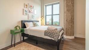 Un dormitorio con una cama, una mesita de noche verde, un ventanal grande y una obra de arte en la pared.