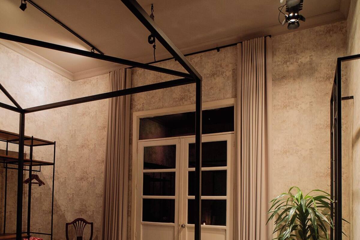 A bedroom with a four-poster bed, a nightstand, a chair, a potted plant, and a large window with curtains.