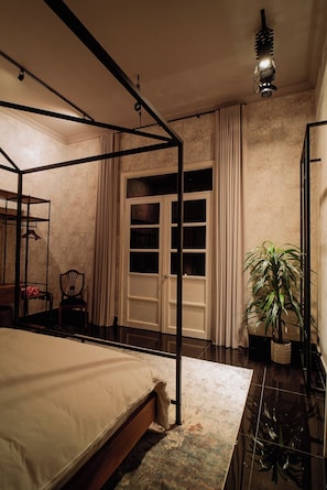 A bedroom with a four-poster bed, a nightstand, a chair, a potted plant, and a large window with curtains. - Hush Hush (Barranquilla)