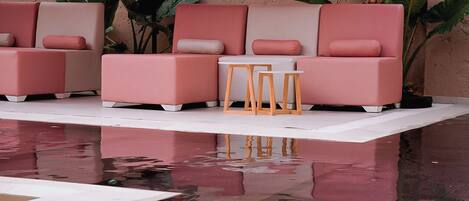 A poolside seating area with pink sofas and two wooden tables, surrounded by large red umbrellas.