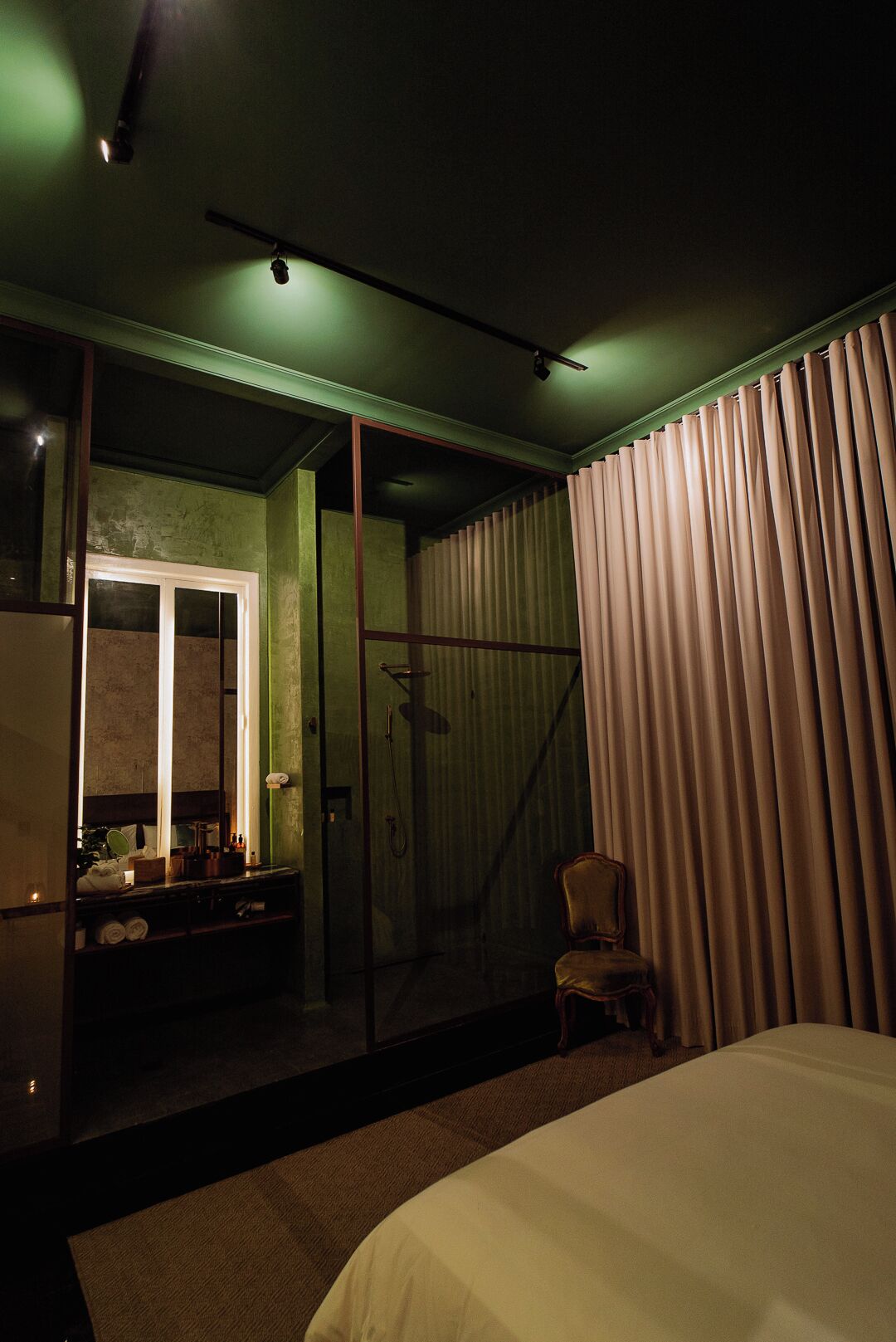 A dimly lit hotel room with a bed, a chair, a bathroom with a mirror, and a curtain.