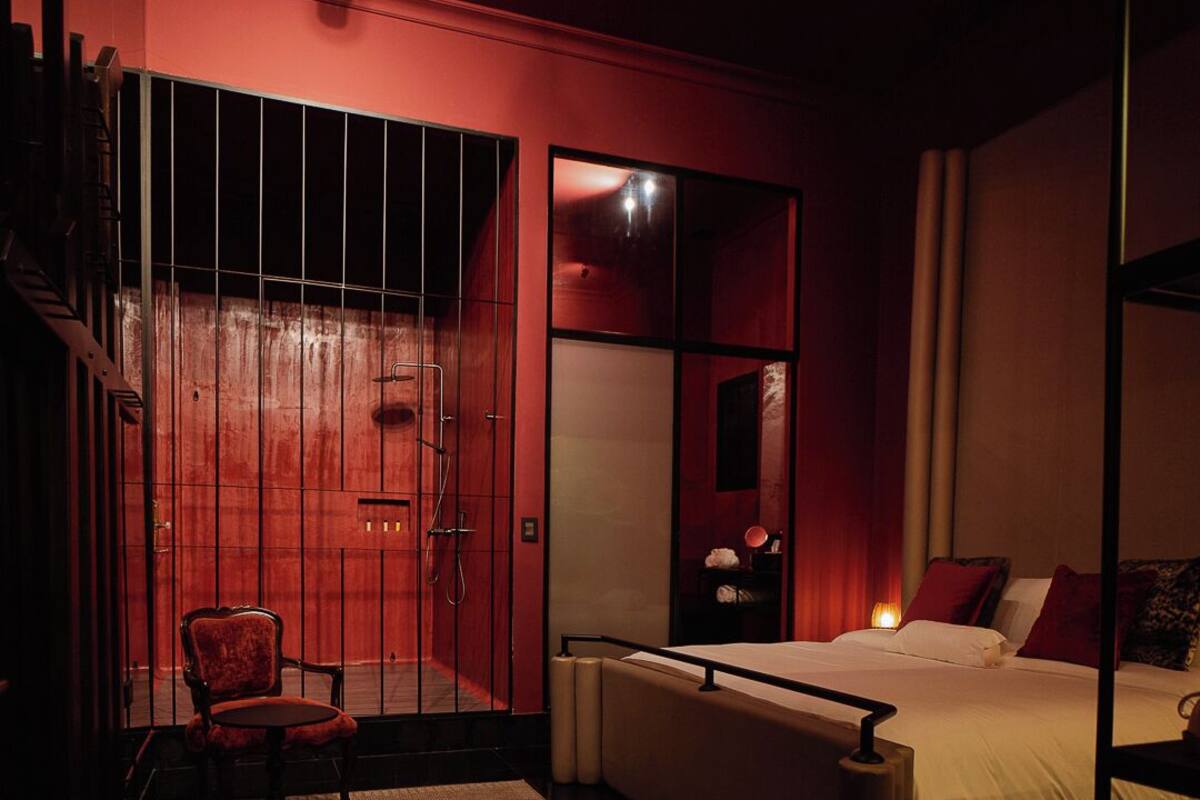 A dimly lit bedroom with a red accent wall, a single bed with white bedding, a chair, and a small table.
