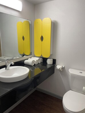 Un baño con lavamanos blanco, espejo e inodoro. Hay armarios amarillos sobre el lavamanos y una encimera negra.