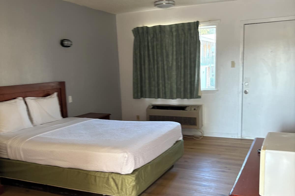 A bedroom with a wooden floor, a bed with a green bedspread, a window with curtains, and a white door.