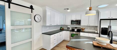 A modern kitchen with white cabinets, a stainless steel refrigerator, a microwave, and a coffee maker. There is a wooden dining table with two chairs.