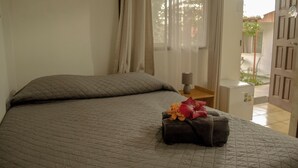 Una cama bien tendida con una colcha acolchada y una almohada decorativa. En la esquina, se ve una mesita con una lámpara y un objeto decorativo.