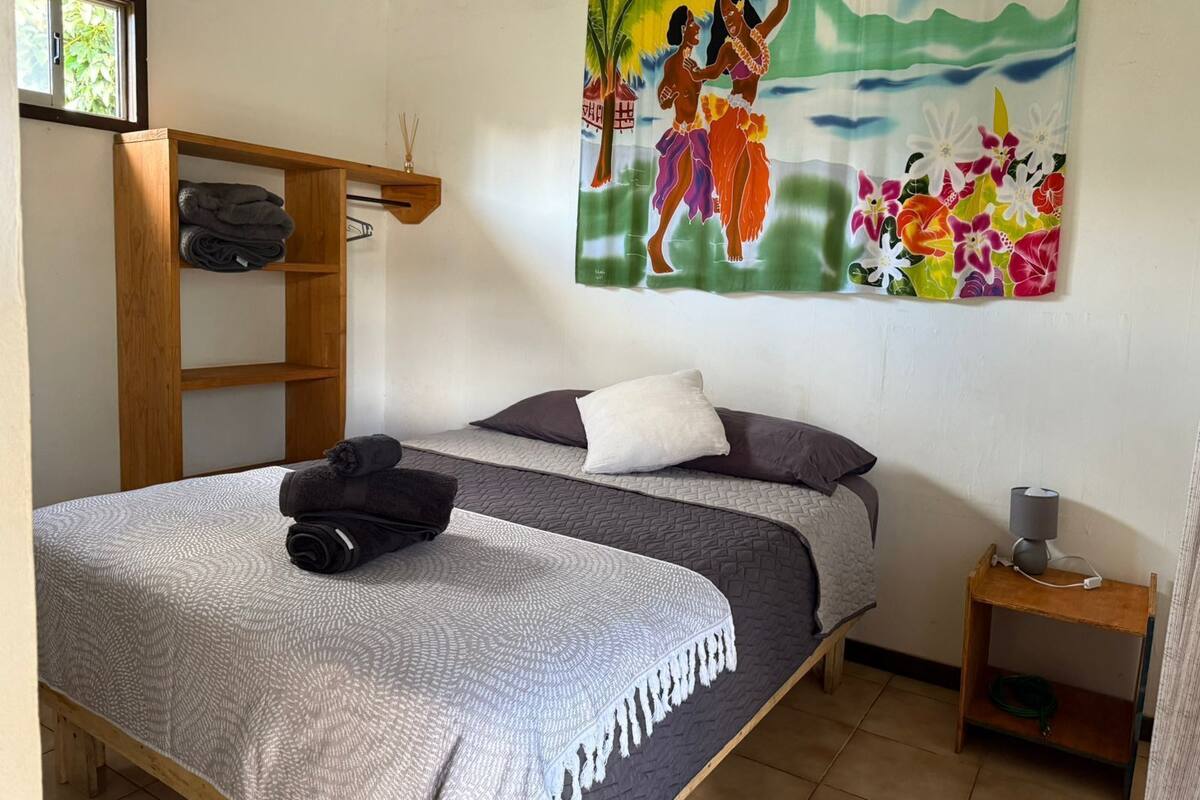 A bedroom with a bed, a wooden shelf, a painting, and a nightstand with a lamp.
