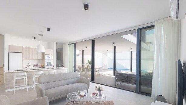 A modern living room with a sofa, coffee table, and TV. A kitchen is visible in the background with a dining area and large windows. - Villa Mae, Brand New, Stunning 4BDR Protaras Villa with Private Pool (Paralimni)
