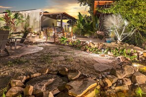 A well-lit outdoor patio area with a wooden pergola, seating, and a small waterfall feature. - Cozy Waterfront Retreat | Pool • Firepit • Sleeps 12 (Bradenton)