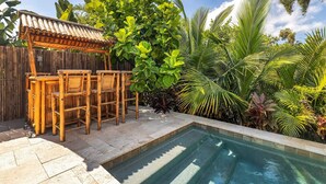 Un área junto a la piscina con una pérgola de bambú y sillas, rodeada de exuberante follaje tropical.