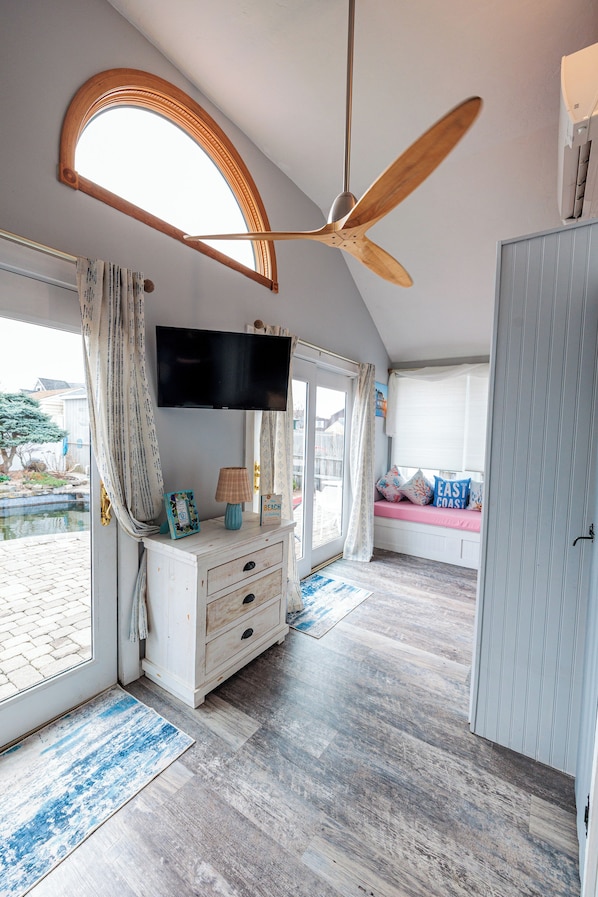 A room with a ceiling fan, a TV mounted on the wall, a window seat with cushions, and a wooden floor.